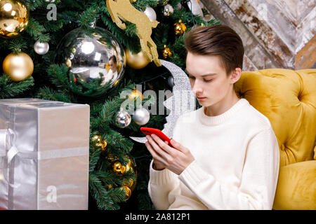 Adolescent garçon assis seul sur un plancher en sapin de Noël avec des cadeaux s'est concentré uniquement sur son smartphone messaging ou contrôler les médias sociaux Smombie - maison de vacances Banque D'Images