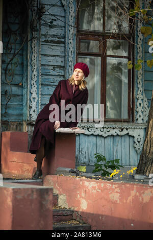 Femme dans un manteau et beret assis sur le perron d'une vieille maison en bois. Banque D'Images