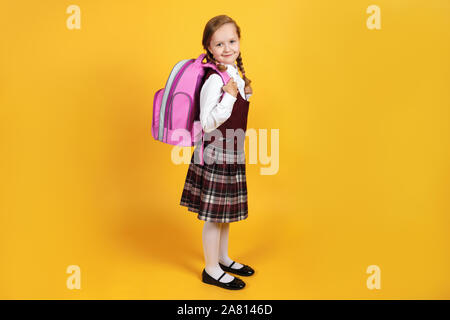 Une vraie lycéenne en uniforme est debout avec un sac à dos derrière son dos sur un fond jaune. Cute little girl est souriant et regardant le Banque D'Images