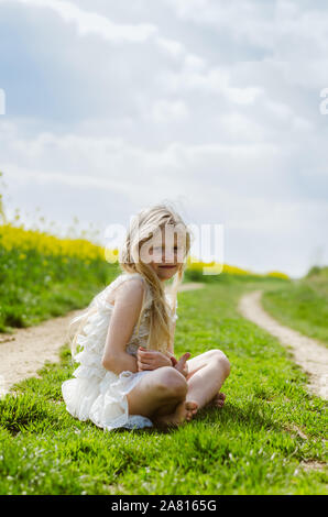 Adorable petite fille blonde aux cheveux longs assis en vert chemin rural Banque D'Images