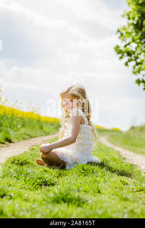 Adorable petite fille blonde aux cheveux longs assis en vert chemin rural Banque D'Images