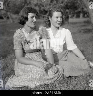 Années 1950, historique, deux jeunes femmes assises ensemble dehors dans un champ, près l'une à côté de l'autre pour leur photo, Angleterre, Royaume-Uni. Banque D'Images