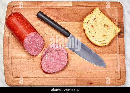 Sur une grande planche à découper en bois se trouve une saucisse coupée en tranches, un morceau de pain blanc, et un couteau de cuisine. Banque D'Images