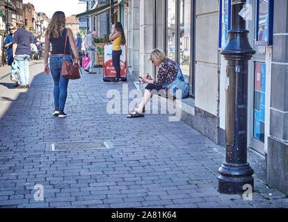 Une femme était assise sur le cill de une vitrine à l'aide de son téléphone mobile comme une autre femme passe devant à Blenheim Street, Belfast, Royaume-Uni Banque D'Images