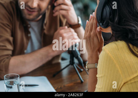 Vue partielle de l'animateur de radio couvrant le microphone avec la main tout en s'asseyant à côté de whispering collègue Banque D'Images