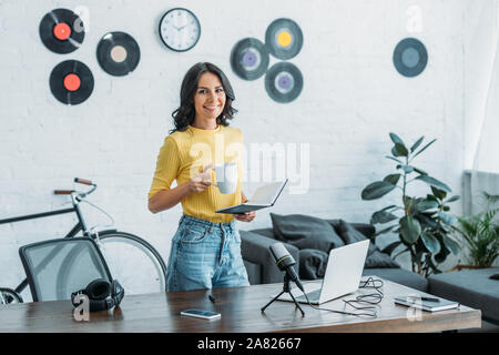 Belle radio host smiling at camera tout en tenant la tasse de café et d'ordinateur portable Banque D'Images