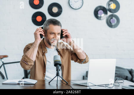 Handsome radio host looking at laptop tout en restant assis sur le lieu de travail et de mettre le casque Banque D'Images