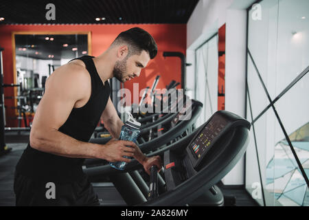 Beau jeune sportif à la recherche sur le tableau de contrôle d'un tapis roulant tout en tenant la bouteille de sport Banque D'Images