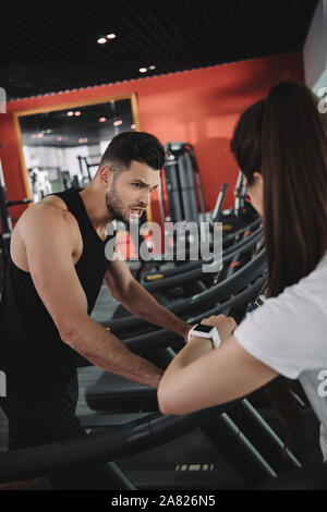 Entraîneur personnel à la remise en forme au tracker en étant debout près de sportsman s'exécutant sur un tapis roulant Banque D'Images