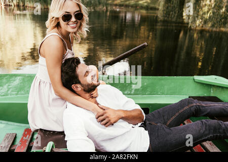 Jolie jeune fille à lunettes hugging heureux petit ami alors qu'il était assis dans un bateau sur le lac Banque D'Images