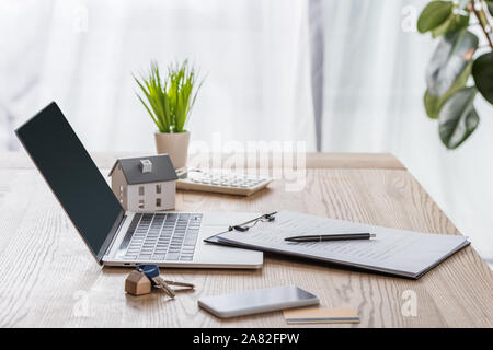 Bureau en bois avec ordinateur portable, smartphone, clés de maison, presse-papiers avec stylo, modèle maison et plante verte Banque D'Images