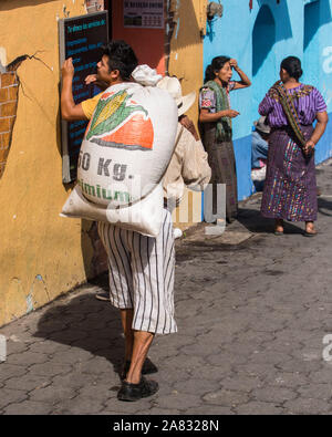 Un vieux homme maya en vêtements traditionnels porte un sac de 50 kg de maïs sur son dos à l'aide d'un portage. Deux femmes en costume traditionnel talk sur le côté de Banque D'Images