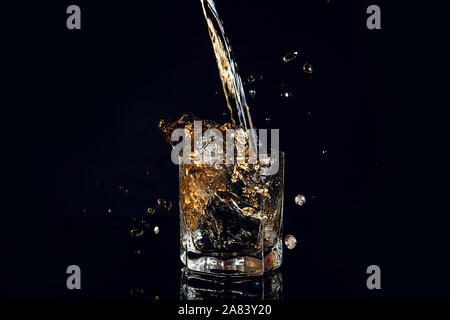 Verser dans un verre à whisky avec de la glace réfléchissante isolé sur la surface noire. Splash de whisky en verre, de nombreuses gouttes de boissons de verre. Banque D'Images