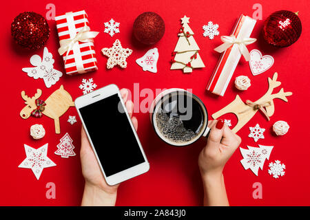 Vue de dessus d'une femme tenant un téléphone dans une main et une tasse de café dans l'autre main sur fond rouge. Décorations de Noël et les jouets. Nouvelle Année holida Banque D'Images