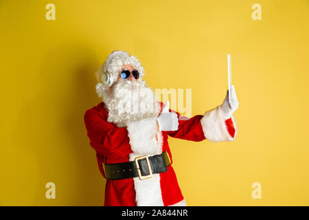 Père Noël avec les gadgets modernes jaune isolé sur fond de studio. Modèle homme de race blanche en costume traditionnel. Concept de Noël, Nouvel An, hiver, humeur technologies. Décisions ou selfies vlog. Banque D'Images