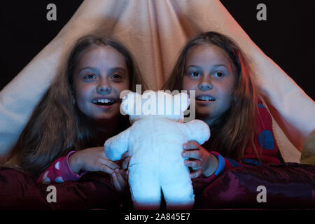 Kids wearing red pyjama au lit sur fond noir. Les amis s'amusant avec des jouets dans une tente. PJs partie pour les enfants. Les enfants et les temps d'amusement concept. Les filles avec des visages heureux de regarder la télévision. Banque D'Images