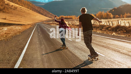 Jeune couple est à cheval sur les longboards par tout droit route de montagne à la lumière au coucher du soleil Banque D'Images