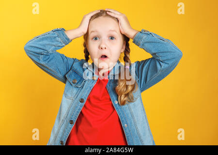 Funny little girl détient les mains derrière sa tête sur un fond jaune. L'enfant est surpris, étonné, émotionnel. Banque D'Images