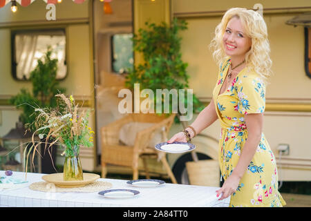 Heureux fille blonde en robe d'été jaune prend les assiettes sur la table et commence à préparer le déjeuner en arrière-plan de camping van remorque. Banque D'Images