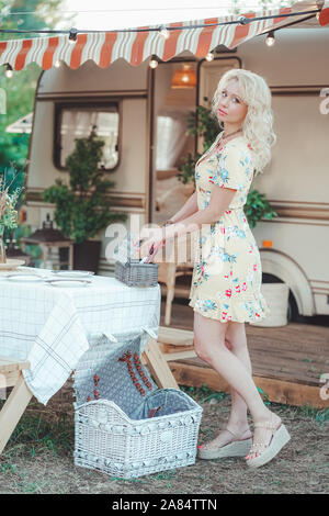 Heureux fille blonde en robe d'été jaune prend les assiettes sur la table et commence à préparer le déjeuner en arrière-plan de camping van remorque. Banque D'Images