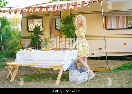 Heureux fille blonde en robe d'été jaune prend les assiettes sur la table et commence à préparer le déjeuner en arrière-plan de camping van remorque. Banque D'Images