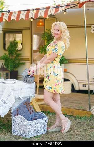 Heureux fille blonde en robe d'été jaune prend les assiettes sur la table et commence à préparer le déjeuner en arrière-plan de camping van remorque. Banque D'Images