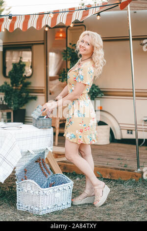 Heureux fille blonde en robe d'été jaune prend les assiettes sur la table et commence à préparer le déjeuner en arrière-plan de camping van remorque. Banque D'Images