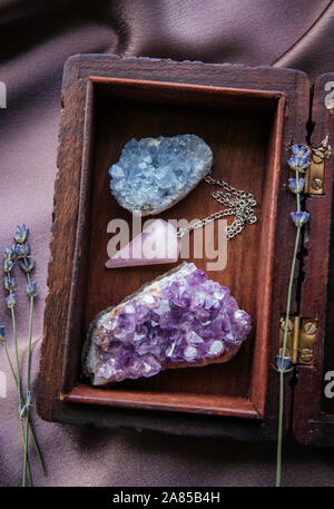 Sorcière outils à l'intérieur belle ancienne boîte en bois. Pendule à quartz rose, amas de cristaux d'améthyste naturel et de célestite. Fleurs de lavande sèches sur violet foncé Banque D'Images