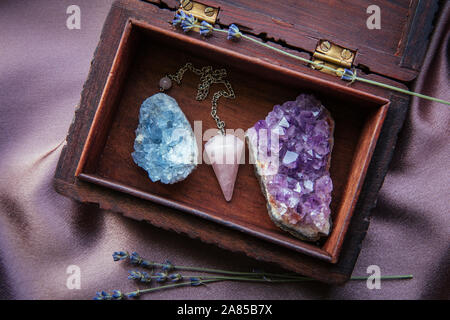 Sorcière outils à l'intérieur belle ancienne boîte en bois. Pendule à quartz rose, amas de cristaux d'améthyste naturel et de célestite. Fleurs de lavande sèches sur violet foncé Banque D'Images