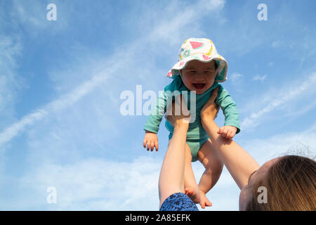 Mother holding baby girl up dans l'air avec les deux bras contre un ciel bleu Banque D'Images