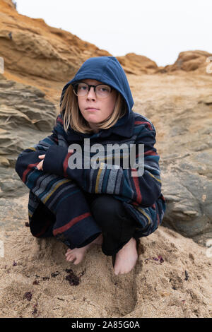 Le Tween crouching in sand on beach looking at camera wearing glasses Banque D'Images