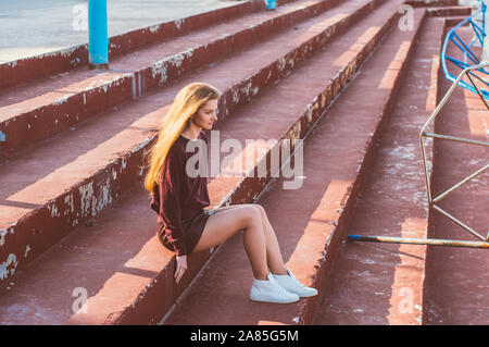 Jeune fille blonde en jupe assise sur un escalier peint de bâtiment abandonné Banque D'Images