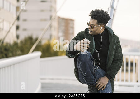 Beau jeune homme noir africain avec des lunettes à l'écoute de la musique sur smartphone avec écouteurs en position assise, dans la ville, le mode de vie et la technologie conc Banque D'Images