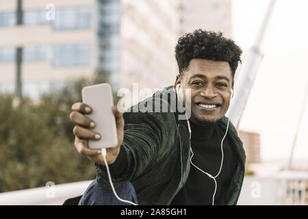 Beau jeune homme noir africain faisant un ou selfies un appel vidéo avec le smartphone et d'écouteurs en position assise, dans la ville, le mode de vie et de techno Banque D'Images