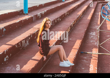 Jeune fille blonde en jupe assise sur un escalier peint de bâtiment abandonné Banque D'Images