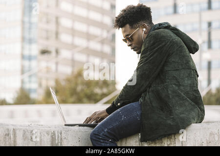 Beau jeune étudiant avec des lunettes et d'écouteurs working on laptop assis à l'extérieur dans la ville, le mode de vie et la technologie à l'aide d'internet concept e Banque D'Images