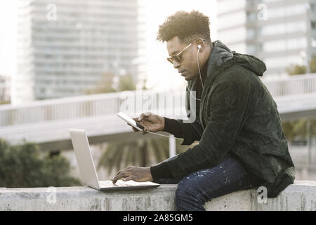 Beau jeune étudiant avec des lunettes et d'écouteurs travailler avec ordinateur portable et téléphone assis à l'extérieur de la ville au coucher du soleil, le mode de vie et de la technologie Banque D'Images