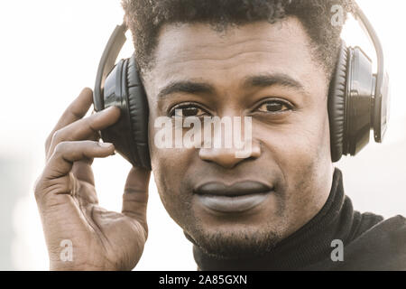 Portrait of a happy african black man smiling écouter la musique avec une main en contact avec le casque au coucher du soleil, de la technologie et le style de concept Banque D'Images