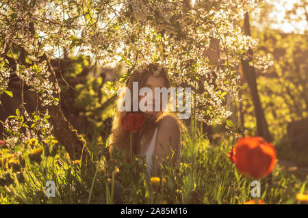 Fille en robe blanche assis parmi les fleurs près de tulip dans le coucher du soleil, les pissenlits et les fleurs de cerisier au printemps Banque D'Images