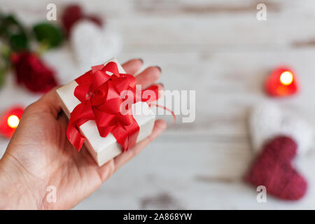 Cadeaux Saint Valentin. Beauty woman hands holding gift box avec arc rouge sur fond de vacances de bougies, de fleurs de rose et des coeurs. Banque D'Images