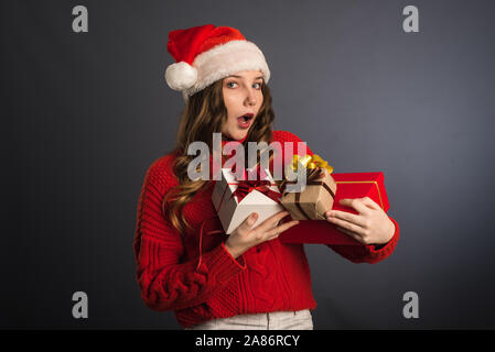 Surpris fille avec un tas de cadeaux dans les mains. Une belle fille avec un visage exprimant l'émotion de wow, dans un chandail rouge et un chapeau de Noël Sa Banque D'Images