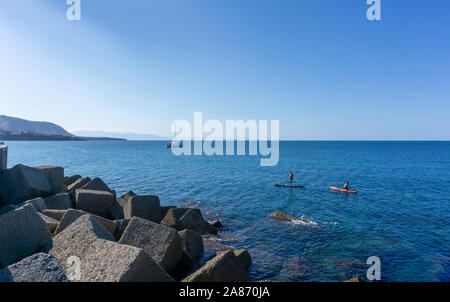 Au large de la côte de Cefalú, Sicile, Italie, deux hommes paddle dans les eaux bleu clair de la méditerranée, avec un yacht au loin. Banque D'Images