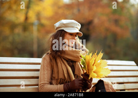 Bonjour l'automne. moyen age femme en pull, jupe, chapeau, gants et écharpe assis sur un banc à l'extérieur dans le parc automne à à feuilles jaunes. Banque D'Images