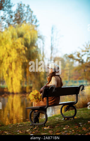 Bonjour l'automne. Full Length portrait of smiling woman in trendy pull, jupe, chapeau, gants et écharpe avec feuilles jaunes à la recherche dans la distance tout en Banque D'Images