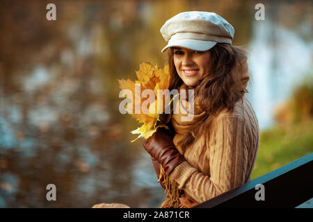 Bonjour l'automne. Portrait of smiling femme moderne en pull, chapeau, gants et écharpe avec feuilles jaunes tout en restant assis sur un banc à l'extérieur dans l'automne pa Banque D'Images