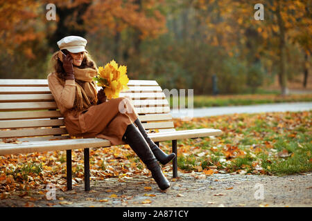 Bonjour l'automne. Portrait de jeune femme en pull, jupe, chapeau, gants et écharpe avec des feuilles jaunes de parler sur un téléphone mobile en position assise Banque D'Images