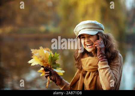 Bonjour l'automne. Portrait of smiling trendy moyen age femme en pull, chapeau, gants et écharpe avec feuilles jaunes s'exprimant sur un smartphone en position assise Banque D'Images