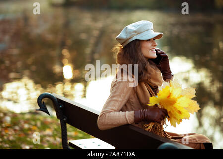 Bonjour l'automne. smiling femme moderne en pull, jupe, chapeau, gants et écharpe avec feuilles jaunes parlant sur un téléphone cellulaire alors qu'il était assis sur un banc outsi Banque D'Images