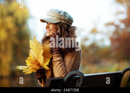 Bonjour l'automne. smiling femme moderne de 40 ans, en pull, chapeau, gants et écharpe avec feuilles jaunes parlant sur un téléphone cellulaire alors qu'il était assis sur un banc Banque D'Images