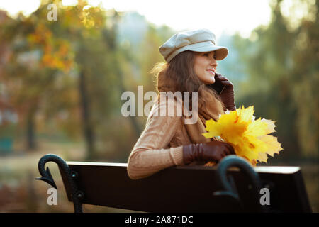 Bonjour l'automne. Heureux femme élégante en pull, chapeau, gants et écharpe avec feuilles jaunes parlant sur un téléphone cellulaire en assise sur un banc à l'extérieur en t Banque D'Images